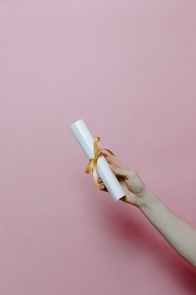 A hand holding a rolled diploma with ribbon on a pink background, symbolizing achievement.